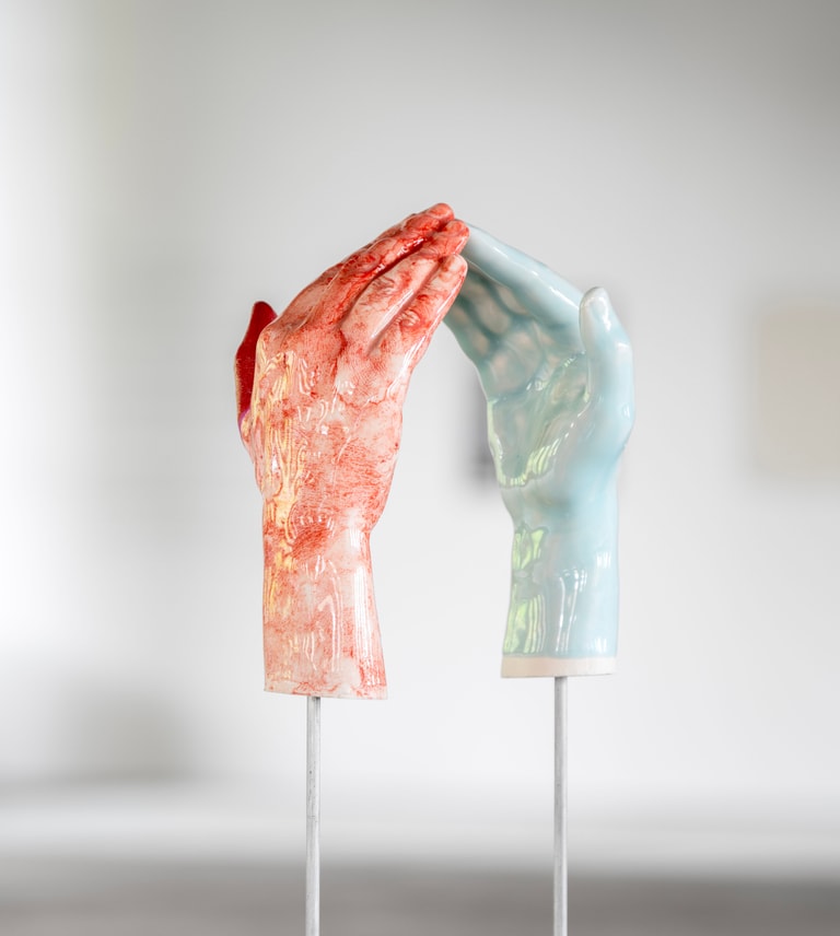 Two ceramic hands, one glazed in red and one in pale blue, suspended on metal rods. Their fingertips almost touch. By Maud Haya-Baviera.